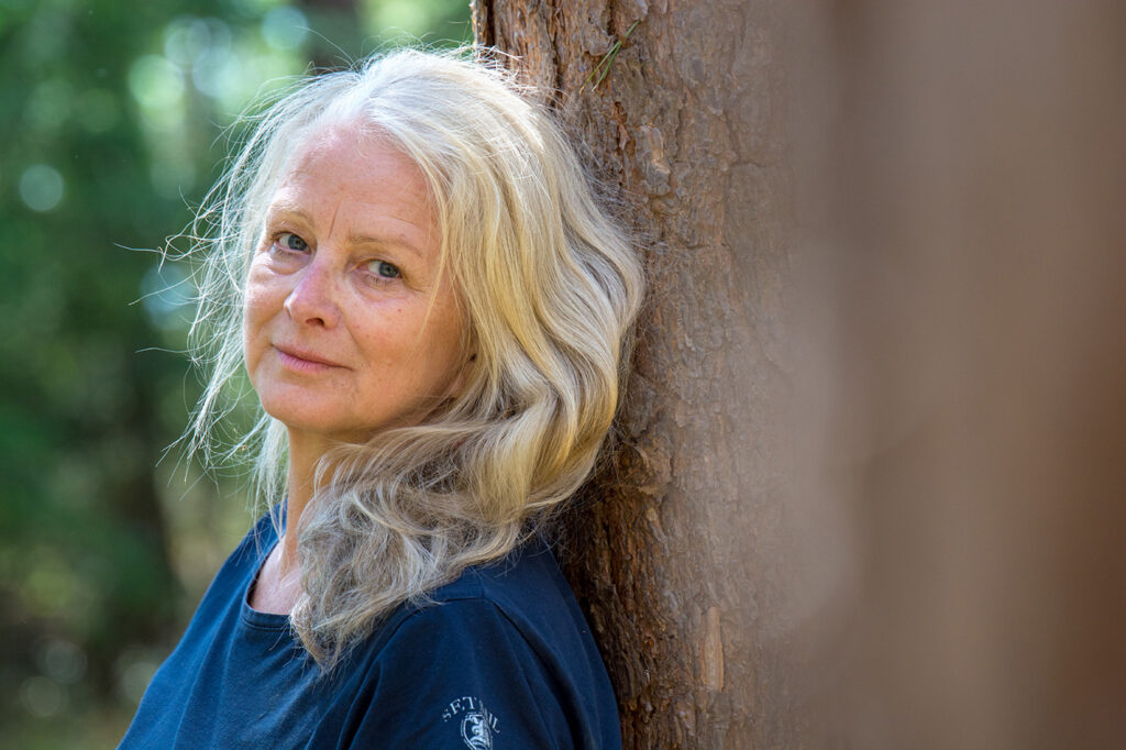 Wunderschöne Frau mit blonden langen Haaren lehnt am Baum und lächelt in die Kamera.
