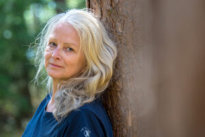 Wunderschöne Frau mit blonden langen Haaren lehnt am Baum und lächelt in die Kamera.
